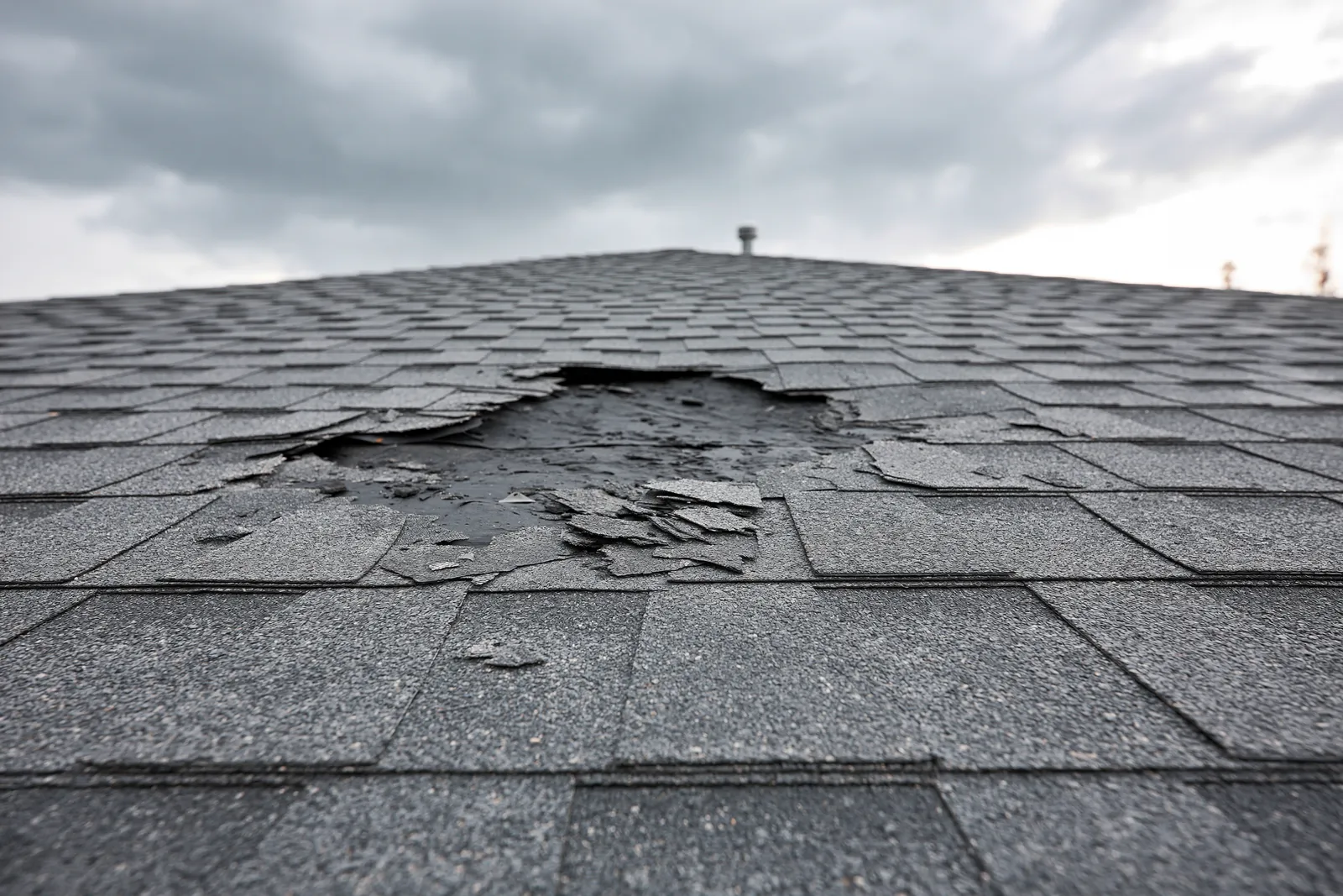 Torn shingles reveal a hole in the roof beneath a stormy sky